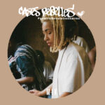 Photo en couleur de profil de Kemistry au carnaval de Notting Hill en 1994. Elle a un t-shirt blanc, des locks blondes coupés au dessus des épaules. Elle regarde vers le bas , ce que l'on imagine être vers ses platines. Une cadre arrondi de couleur marron clair entoure l'image. En haut il est inscrit "Cases Rebelles - PanAfroRévolutionnaires."